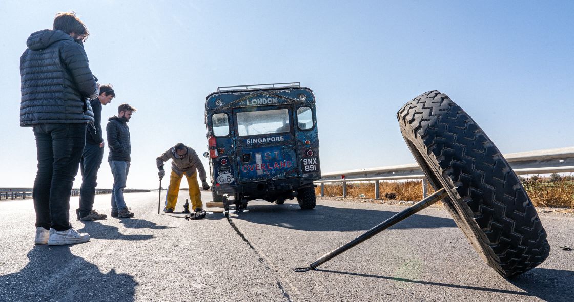 A Group Of People Standing Next To A Vehicle With A Large Tire