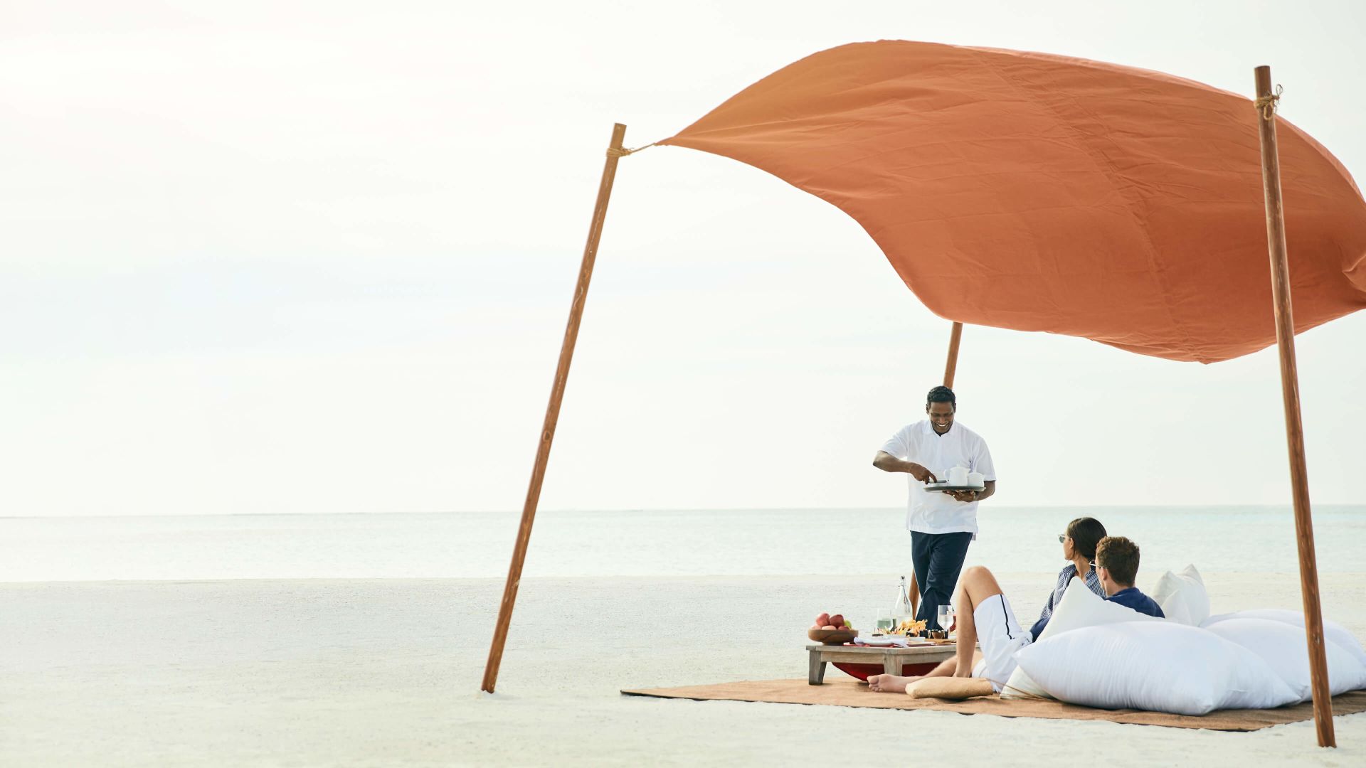 A Man Standing Next To A Couple Of People Sitting Under An Umbrella