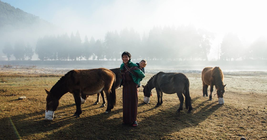 A Person Holding A Baby And Standing Next To A Group Of Horses