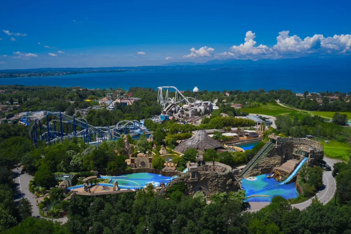 The image shows an amusement park on Lake Garda in Italy. There are various roller coasters and water attractions surrounded by greenery. In the background you can see the lake and the mountains on the horizon. The weather is sunny and the sky is blue with a few clouds.