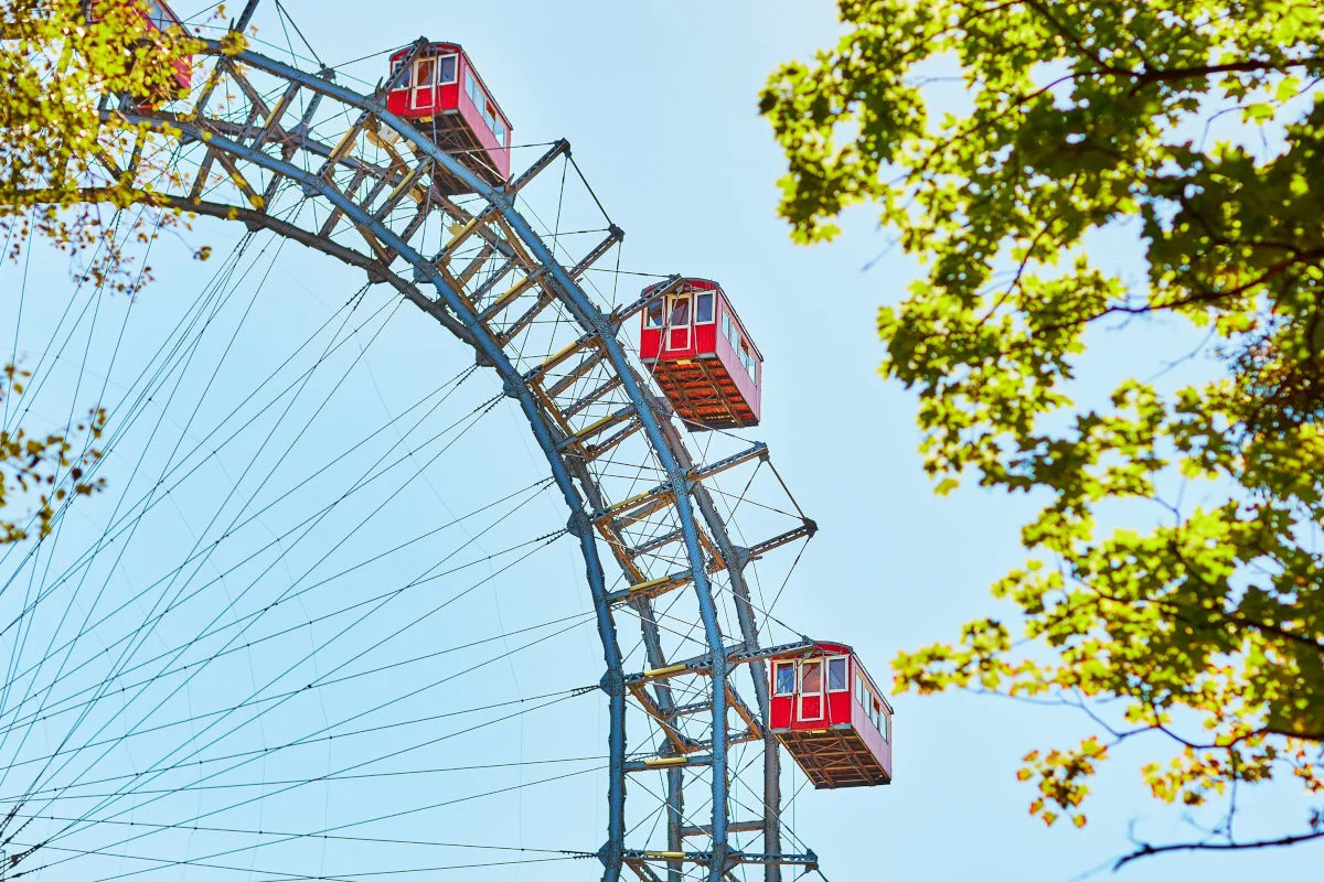 Pictured here is the historic Ferris wheel at the Prater amusement park in Vienna. Red gondolas against a blue sky among green leaves.