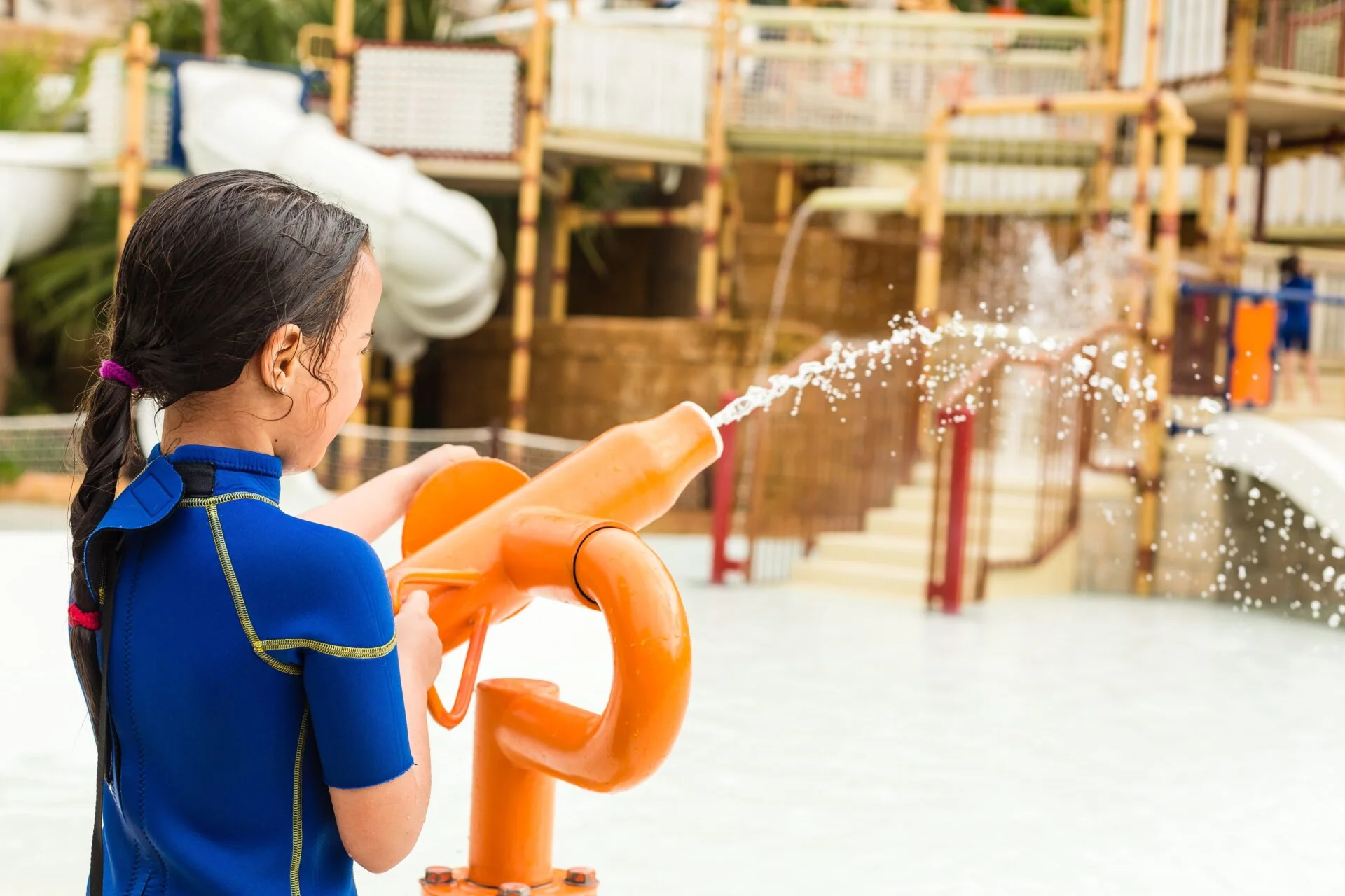 Pictured is a child in a blue bathing suit, playing at the Siam Park water park in Tenerife. The child is holding an orange water cannon from which water is gushing. The water slides and structures of the water park, which are built in a tropical theme, are visible in the background.