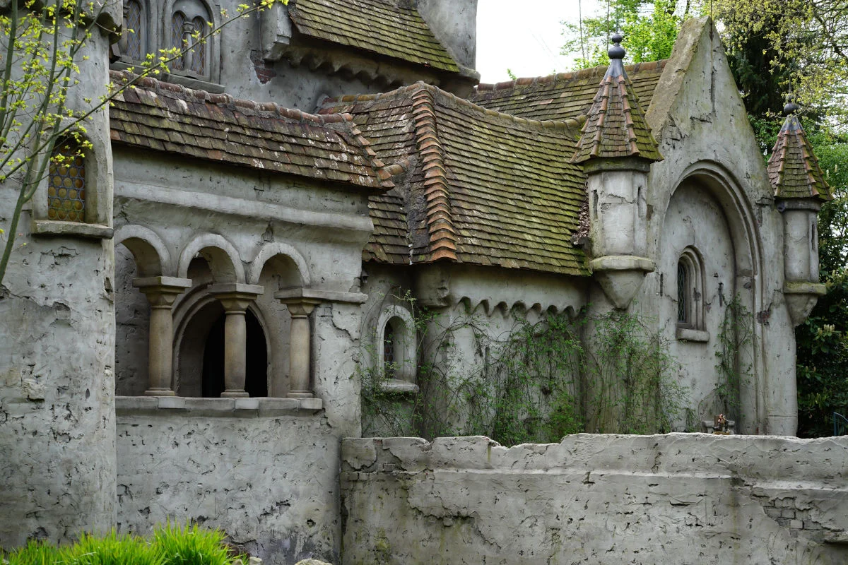 Pictured is an old fairy tale house with arched windows and pointed roofs.It is the match girl's house from the Forest of Fairy Tales.It looks abandoned and there are plants climbing up its walls. The roof is covered in moss, which adds to its mysterious appearance.