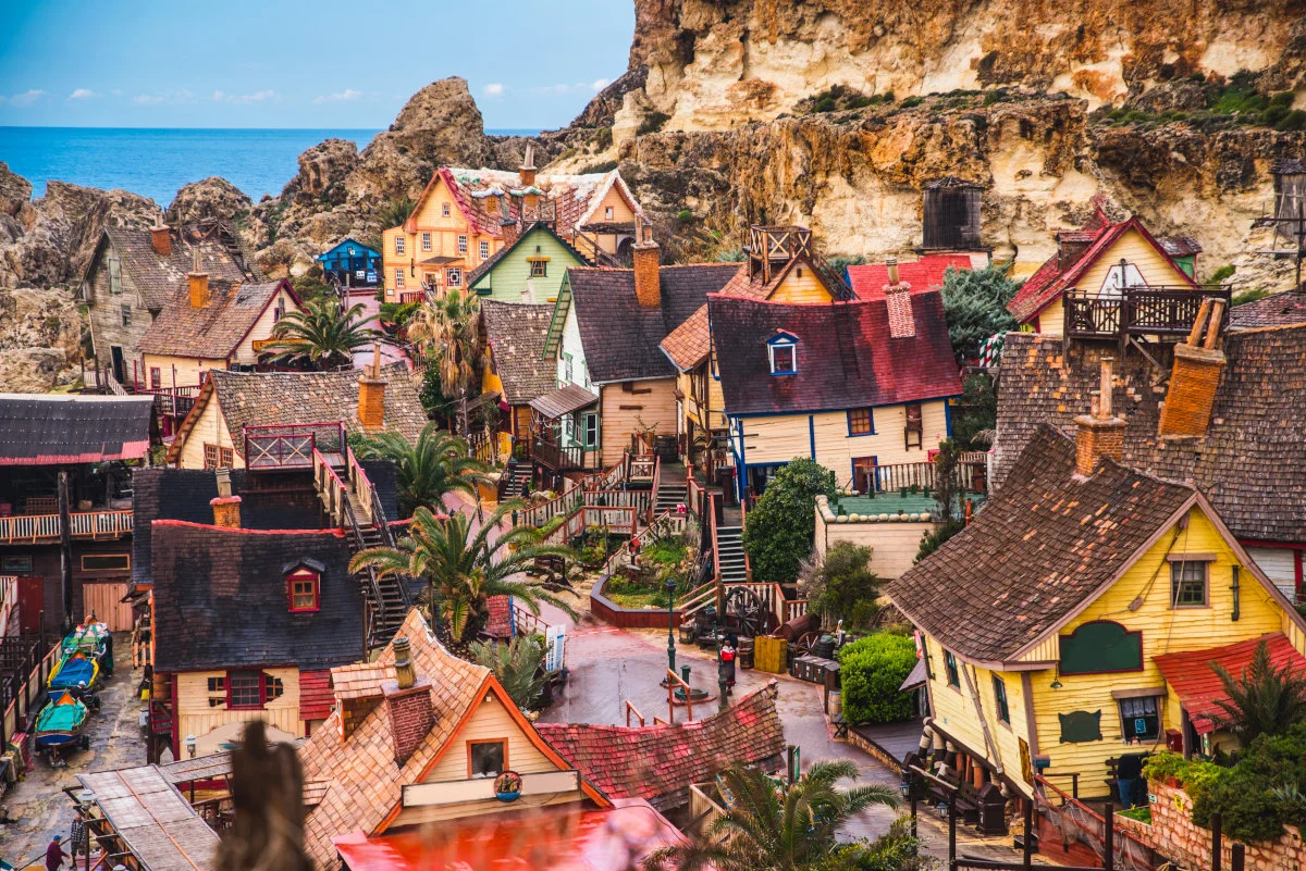 This photo shows Popeye Village in Malta. The colourful cottages are situated near the coast, surrounded by cliffs and palm trees.